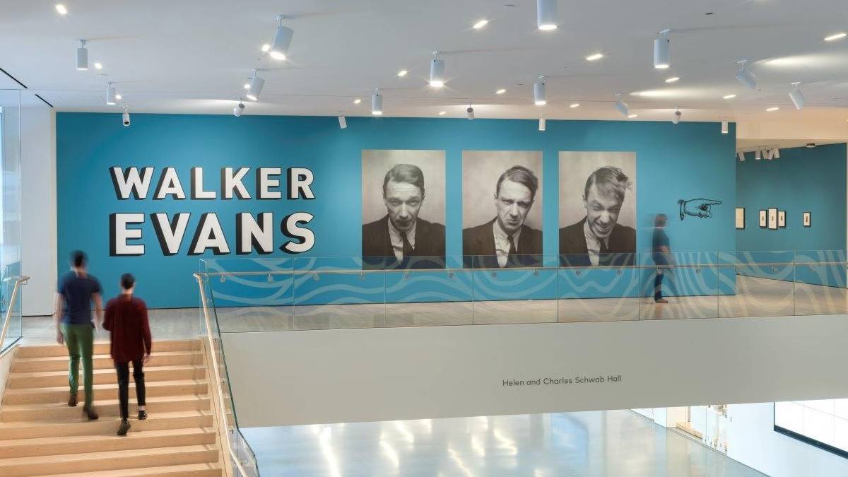 Two people walking up stairs to a landing with a blue wall and three photos of a mans face on the wall